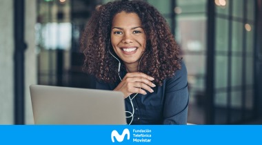 Mujer con cabello abundante, usando audífonos y sonriendo frente a su computador