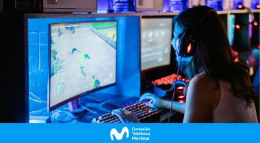 Mujer joven con audífonos frente a una pantalla de computador que refleja una luz azul y gráficos de videojuegos